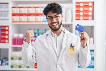 Hispanic man with beard working at pharmacy drugstore holding credit card screaming proud,...