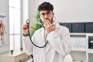 Hispanic man with beard wearing doctor uniform and stethoscope covering mouth with hand, shocked...