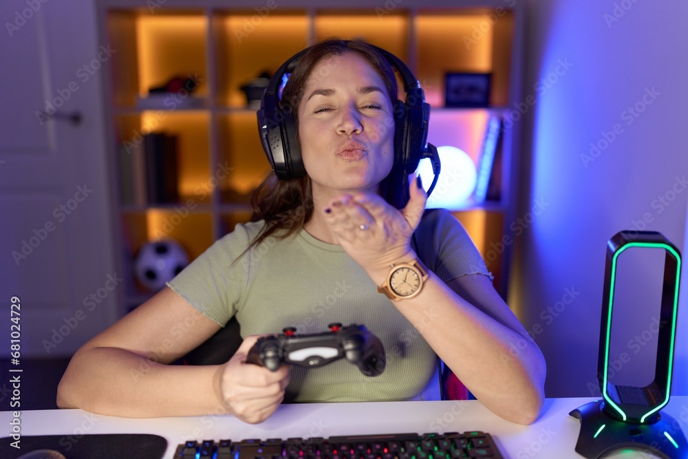 Sticker Beautiful brunette woman playing video games wearing headphones looking at the camera blowing a kiss with hand on air being lovely and sexy. love expression.