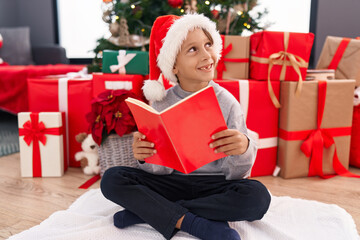 Obraz na płótnie Canvas Adorable hispanic boy reading book sitting on floor by christmas tree at home