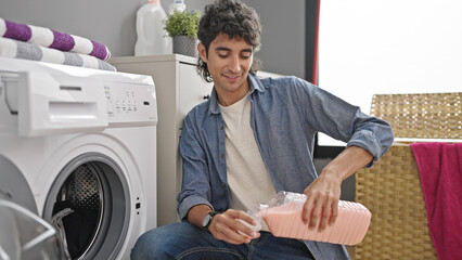 Young hispanic man washing clothes pouring detergent smiling at laundry room