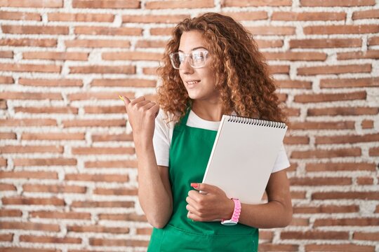 Young caucasian woman holding art notebook smiling with happy face looking and pointing to the side with thumb up.