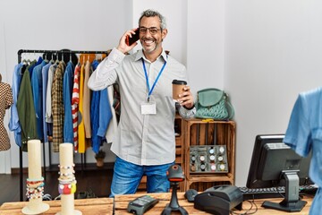 Middle age grey-haired man shop assistant talking on smartphone drinking coffee at clothing store