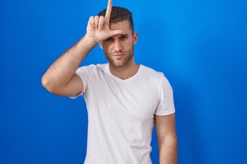 Young caucasian man standing over blue background making fun of people with fingers on forehead...
