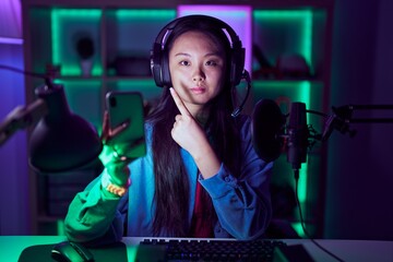 Young asian woman playing video games with smartphone pointing with hand finger to the side showing advertisement, serious and calm face