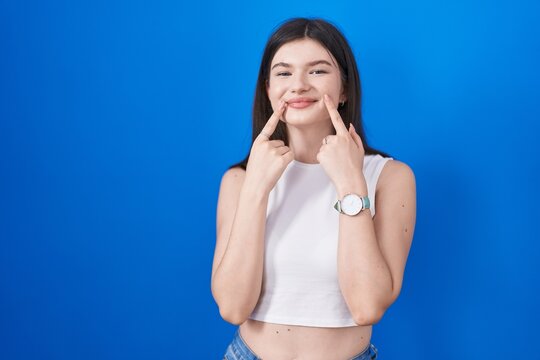 Young caucasian woman standing over blue background smiling with open mouth, fingers pointing and forcing cheerful smile