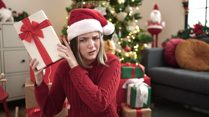 Young blonde woman hearing gift sound sitting on floor by christmas tree at home