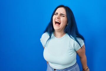 Young modern girl with blue hair standing over blue background angry and mad screaming frustrated and furious, shouting with anger. rage and aggressive concept.