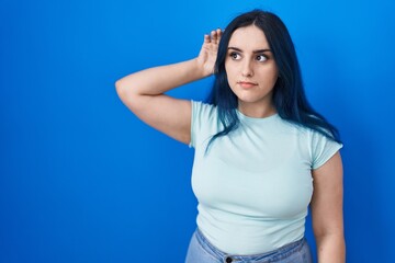 Young modern girl with blue hair standing over blue background smiling with hand over ear listening...