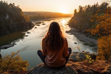 Young woman sits on a stone overlooking the river at sunset and meditates, early autumn