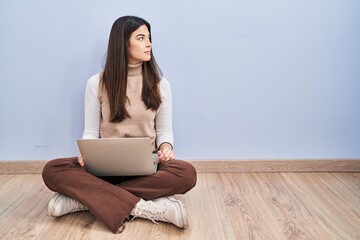 Young brunette woman working using computer laptop sitting on the floor looking to side, relax profile pose with natural face and confident smile.