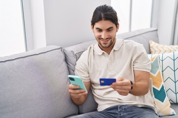 Young hispanic man using smartphone and credit card sitting on sofa at home