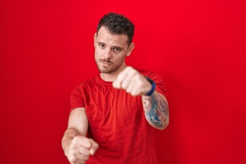 Young hispanic man standing over red background punching fist to fight, aggressive and angry...