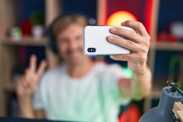 Young blond man streamer smiling confident make selfie by smartphone at gaming room