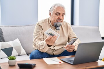 Middle age grey-haired man using laptop counting dollars at home