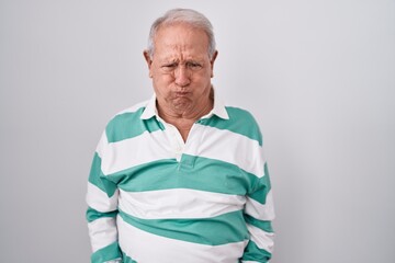 Senior man with grey hair standing over white background puffing cheeks with funny face. mouth...