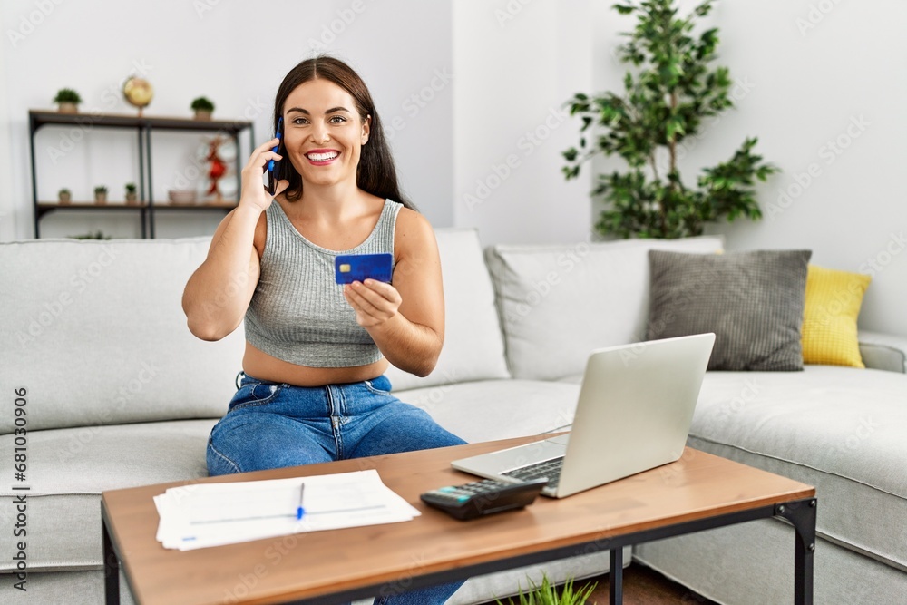 Wall mural Young beautiful hispanic woman using credit card talking on smartphone at home