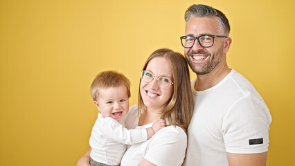 Family of mother, father and baby smiling together looking a the camera over isolated yellow background