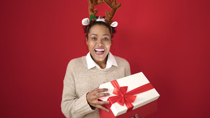 African american woman unpacking christmas gift wearing reindeer ears over isolated red background