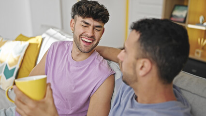 Two men couple drinking coffee sitting on sofa speaking at home