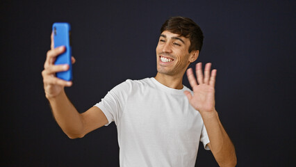 Joyful young hispanic man confidently smiling while standing, talking on phone and having fun during a video call session against a cool isolated black wall background