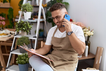 Young hispanic man florist talking on smartphone reading clipboard at flower shop