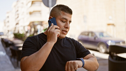 Casual latin man, talking seriously on his smartphone, checks the time on his wristwatch while standing under the sunny urban city outdoors.