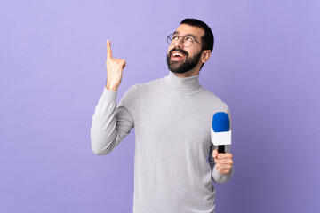 Adult reporter man with beard holding a microphone over isolated purple background pointing up a...