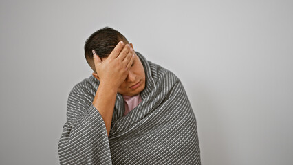 Freezing young latin man wrapped in cozy blanket, braving winter cold indoors, comfort against...