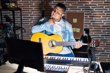 Hispanic young man playing classic guitar at music studio looking confident at the camera smiling with crossed arms and hand raised on chin. thinking positive.