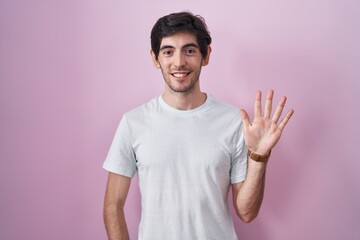 Naklejka premium Young hispanic man standing over pink background showing and pointing up with fingers number five while smiling confident and happy.