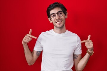 Young hispanic man standing over red background looking confident with smile on face, pointing oneself with fingers proud and happy.