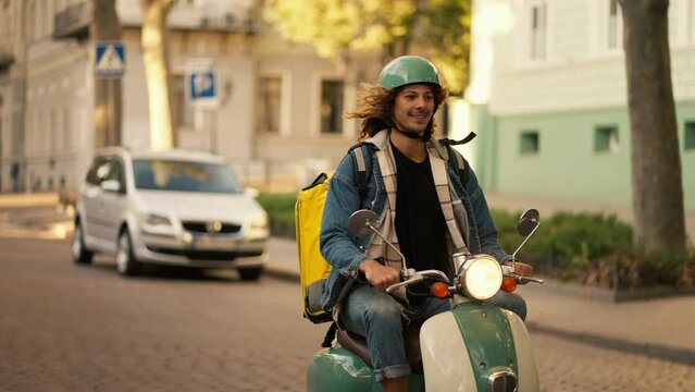 Happy guy with long curly hair in a green helmet and a denim jacket on a green moped with a flashlight on while riding in a summer city