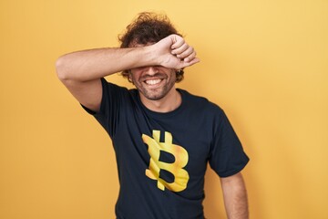 Hispanic young man wearing bitcoin t shirt smiling cheerful playing peek a boo with hands showing...