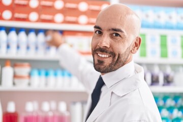 Young bald man pharmacist smiling confident holding bottle of shelving at pharmacy