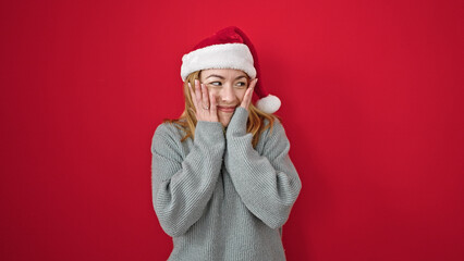 Young blonde woman wearing christmas hat dancing with surprise expression over isolated red background