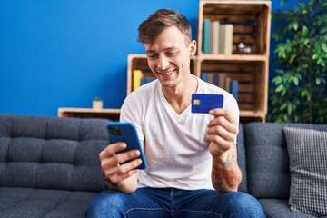 Young man using smartphone and credit card sitting on sofa at home