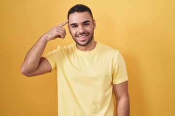Young hispanic man standing over yellow background smiling pointing to head with one finger, great...