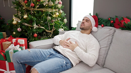 Young hispanic man suffering stomach ache wearing christmas hat at home