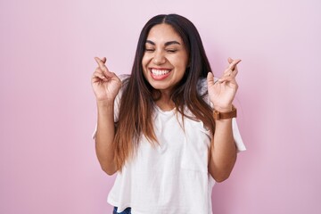 Young arab woman standing over pink background gesturing finger crossed smiling with hope and eyes closed. luck and superstitious concept.