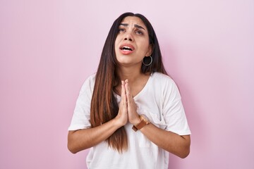Young arab woman standing over pink background begging and praying with hands together with hope...