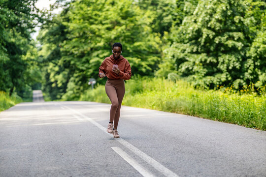 Full body fit active African American woman running fast while exercising outside in nature. Strong athletic black woman sprinting during workout while listening to music with earphones and phone.