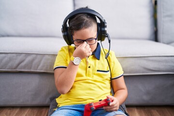 Young hispanic kid playing video game holding controller wearing headphones tired rubbing nose and...