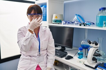 Young brunette woman working at scientist laboratory smelling something stinky and disgusting, intolerable smell, holding breath with fingers on nose. bad smell