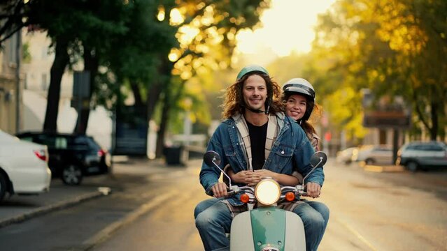 A happy guy in a green moped helmet with long curly hair and a blue denim jacket arrived and stopped with his happy girlfriend in a White helmet on a green moped in a city street on the road in the