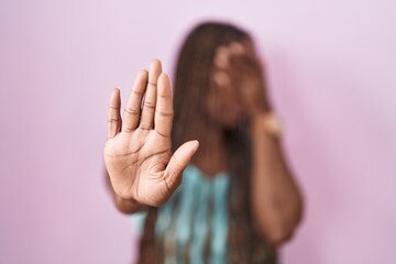 African american woman standing over pink background covering eyes with hands and doing stop...
