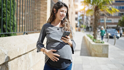 Young pregnant woman using smartphone touching belly at street