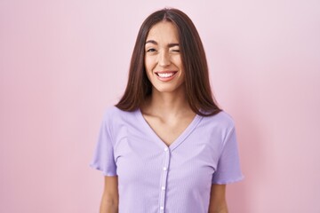 Young hispanic woman with long hair standing over pink background winking looking at the camera...
