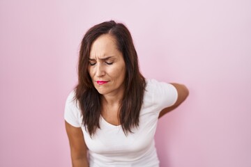 Middle age brunette woman standing over pink background suffering of backache, touching back with hand, muscular pain