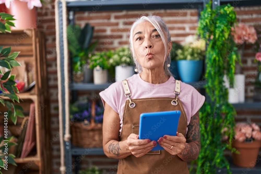 Poster middle age woman with tattoos working at florist shop with tablet looking at the camera blowing a ki